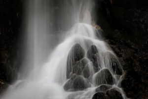 Minimalist black and white fine art photograph of soft flowing waterfall over dark sculpted rocks in a serene natural setting.