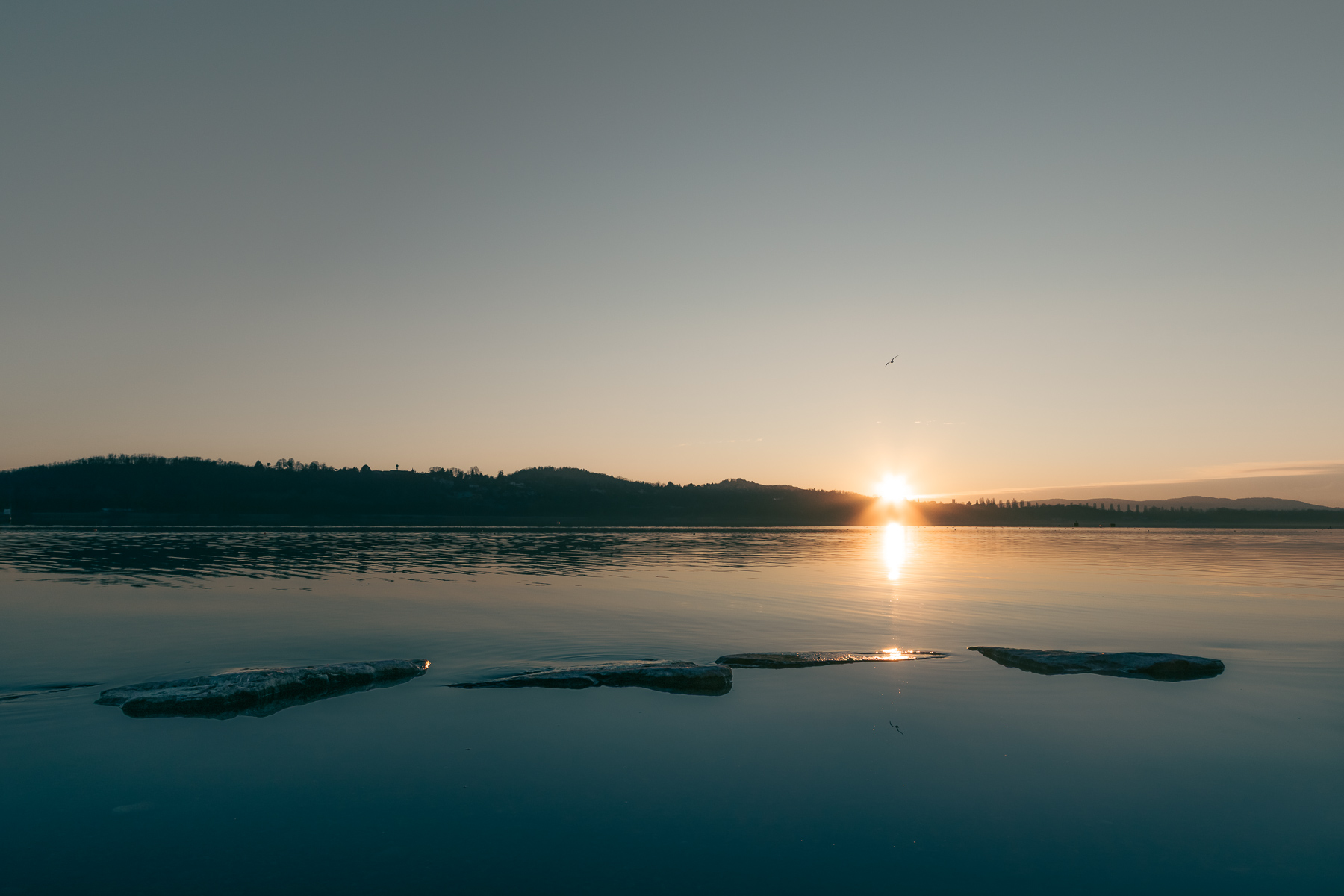 Minimalist lake sunset landscape with stepping stones partially submerged in calm water during golden hour. Warm sunlight reflects across the still lake surface beneath a soft pastel sky, creating a serene fine art nature scene ideal for travel, outdoor, and contemporary wall art photography.
