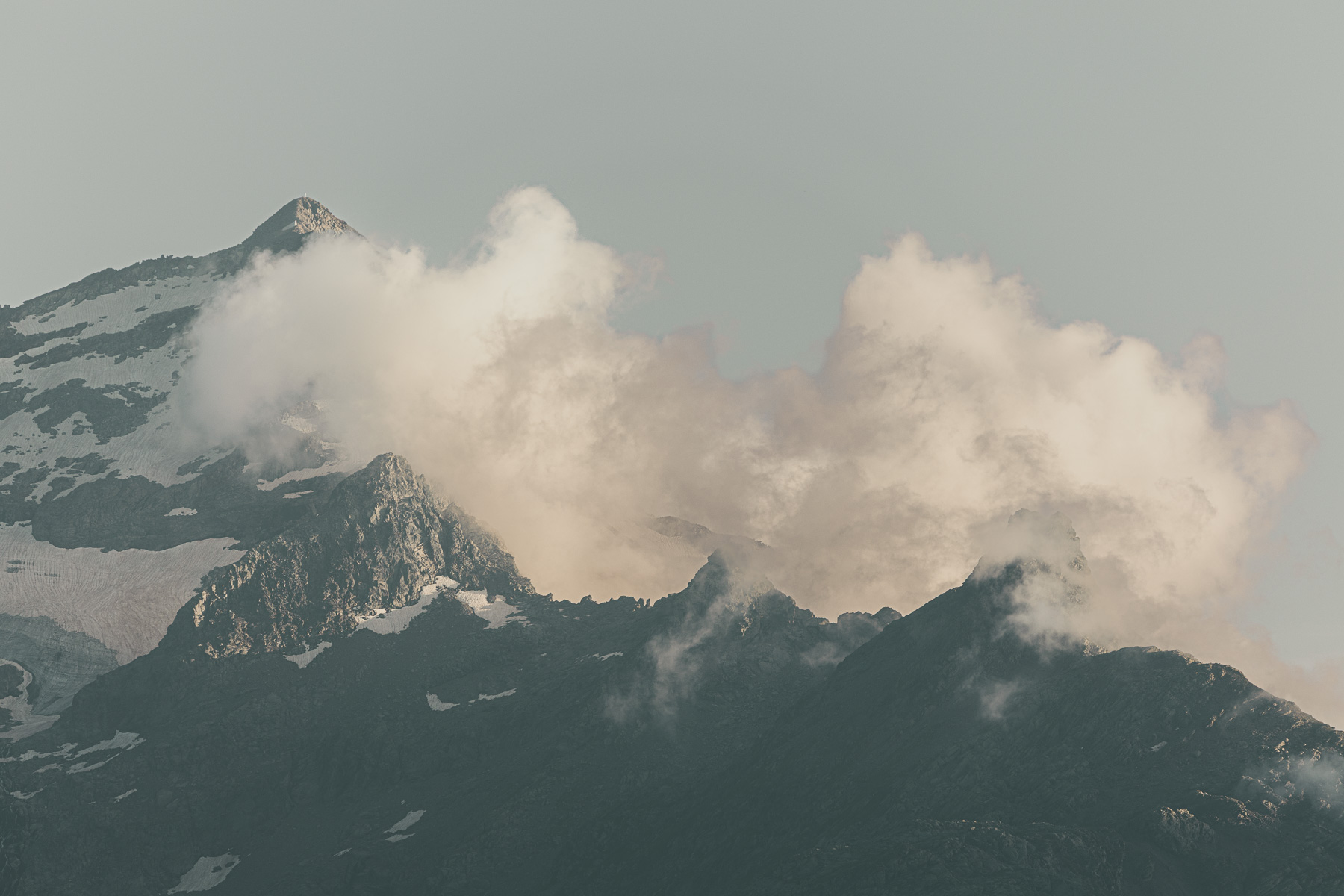 Atmospheric alpine mountain landscape with dramatic clouds drifting over rugged snow-covered peaks during soft golden hour light. Minimalist fine art nature photography featuring textured rocky ridges, muted pastel sky, and cinematic mountain scenery ideal for travel, wilderness, and outdoor journal articles.