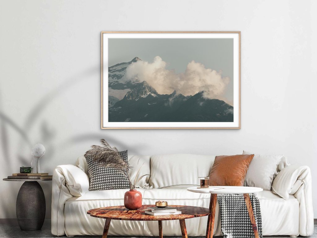 Framed fine art mountain photography print displayed in a modern minimalist living room interior. Atmospheric alpine landscape with dramatic clouds over rugged snow-covered peaks in soft neutral tones, styled as elegant wall art for contemporary home decor and Scandinavian-inspired interiors.