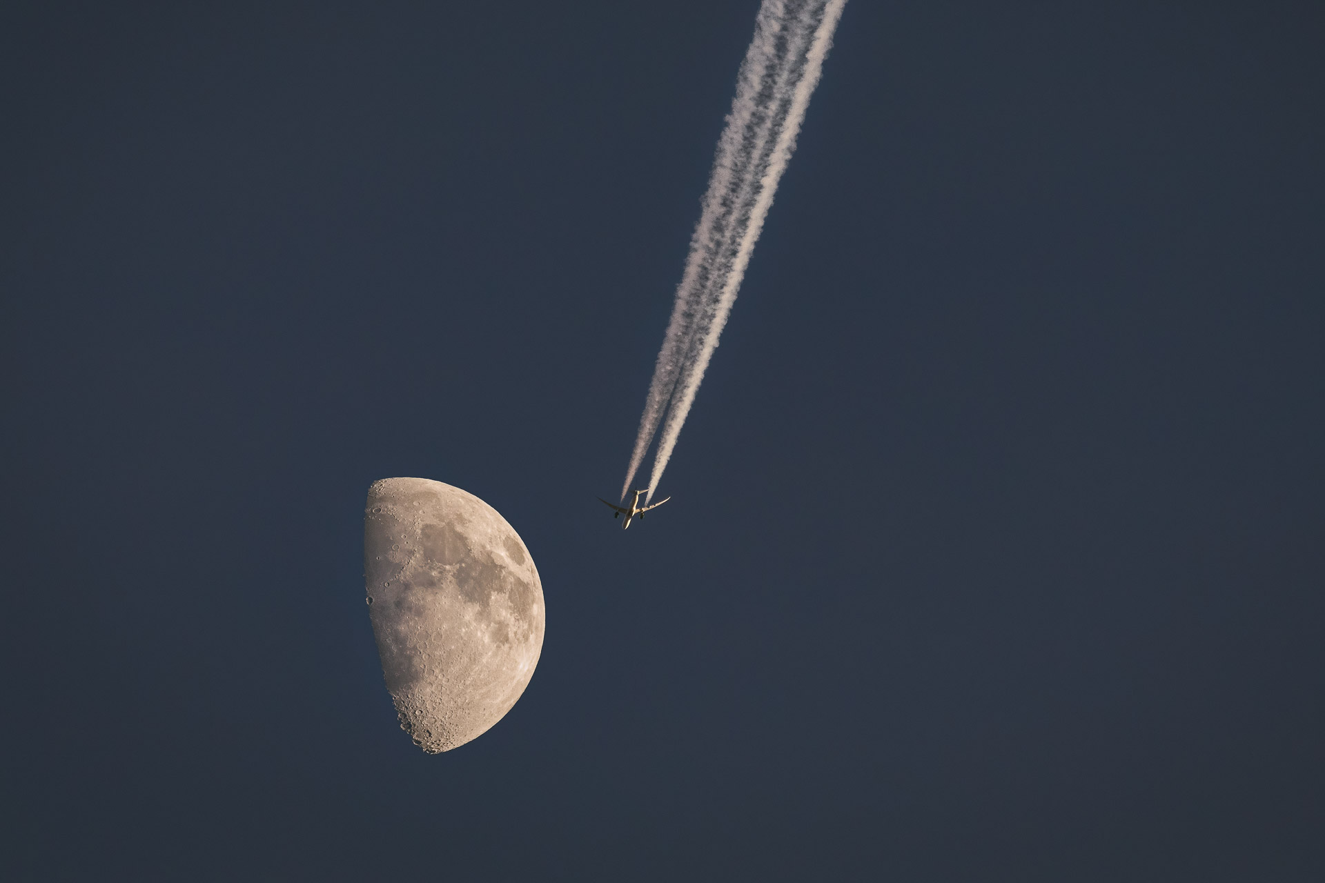Aircraft above and right of a partially illuminated Moon, with two contrails extending diagonally towards the upper edge.