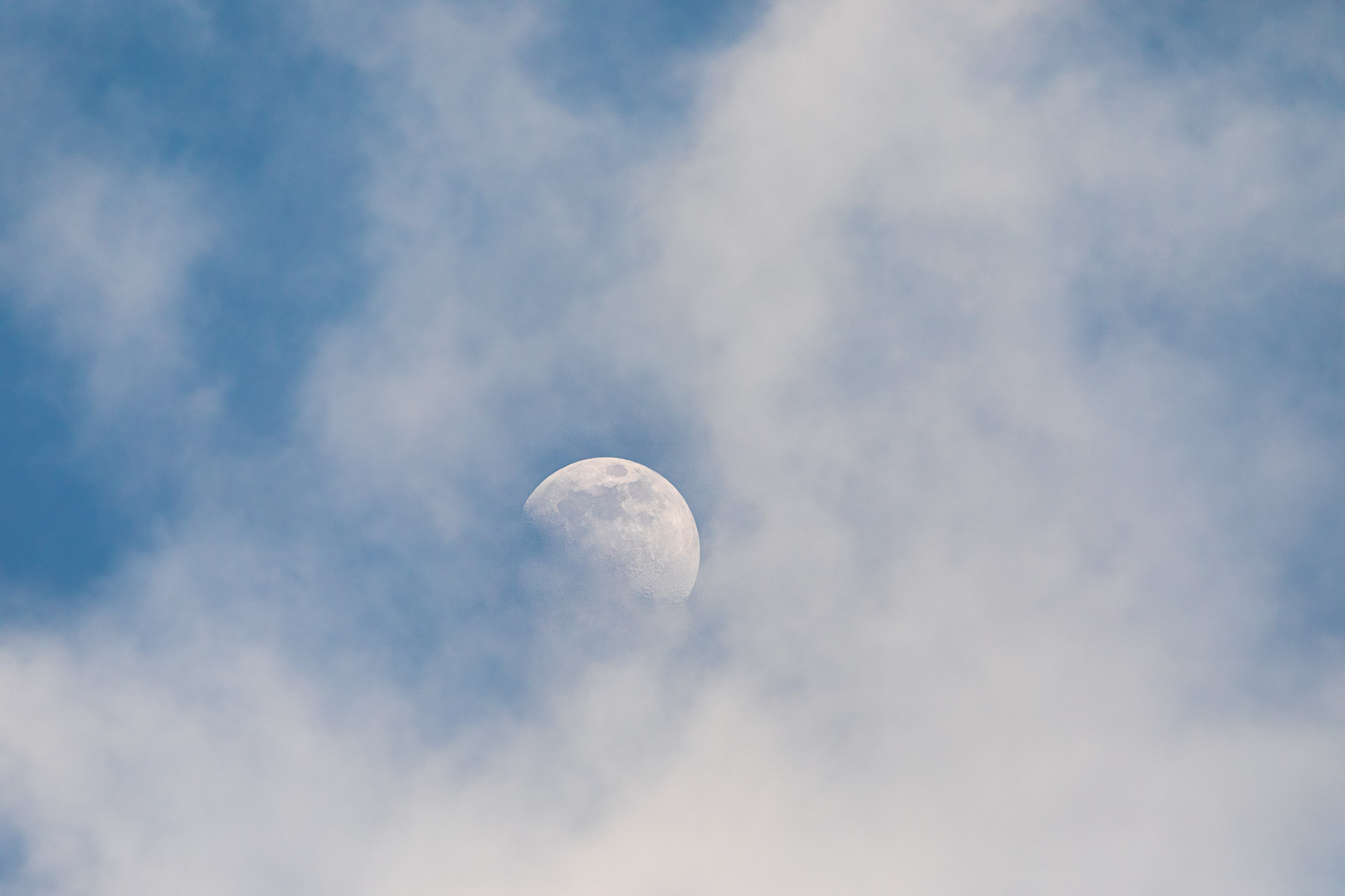 Moon low and slightly left of centre in a blue sky, its outline and surface partly veiled by soft white clouds.